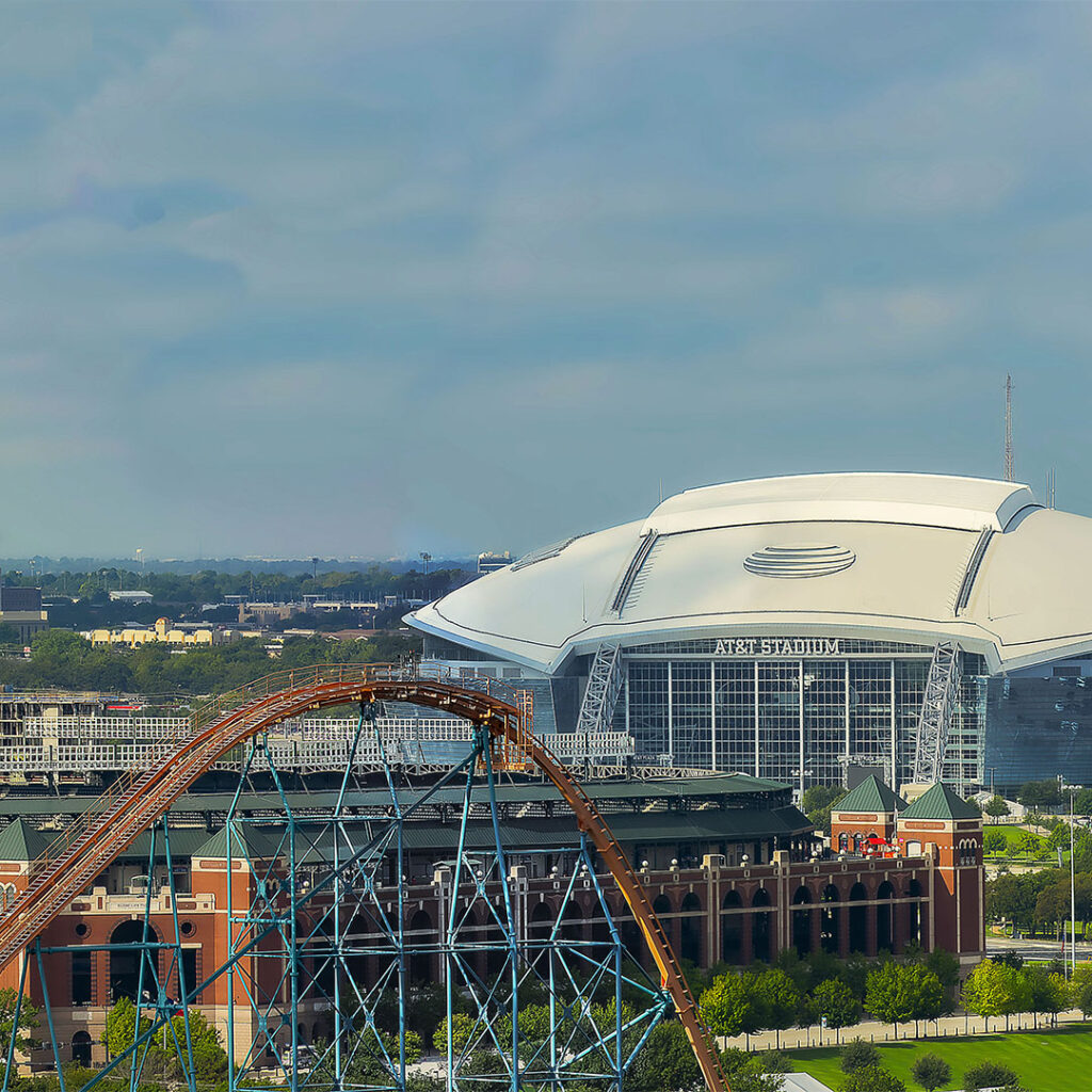 Aerial view of a large white stadium with a retractable roof, surrounded by cityscape and trees. In the foreground, there is a roller coaster, adding an element of amusement park fun to the scene. The sky is partly cloudy.