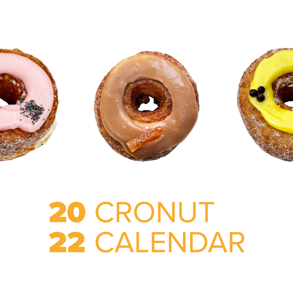 Three colorful cronuts are arranged in a row, each with different toppings: pink with seeds, brown with a candied peel, and yellow with small black spheres. Below, the text reads "2022 CRONUT CALENDAR" in yellow font.