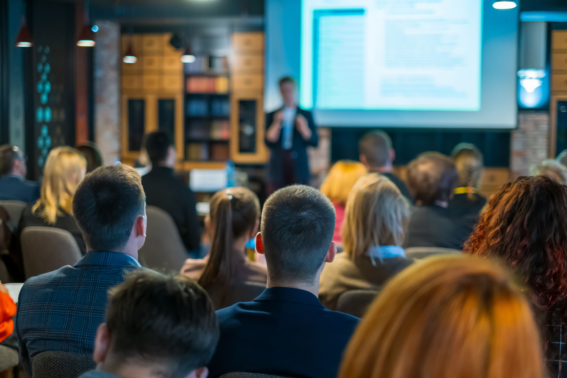 Audience members focus on a speaker presenting at a seminar on web design. The room is dimly lit, and a projector screen displays content at the front. Attendees are seated, facing the presenter, in a classroom or workshop setting.