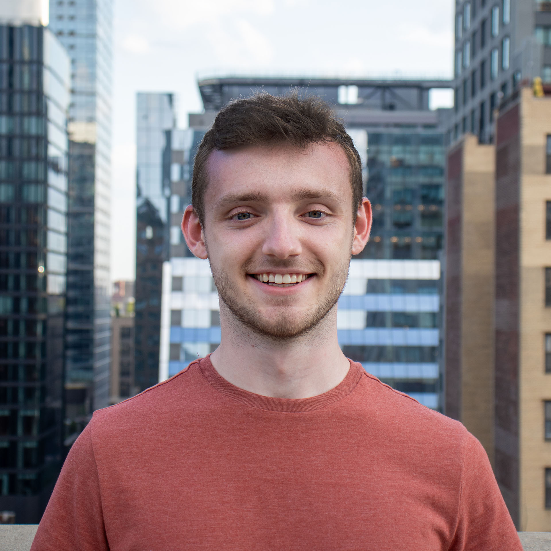 A man in a red shirt smiles at the camera with a cityscape of tall buildings in the background. The sky is clear, and the scene appears to be taken on a rooftop or terrace.