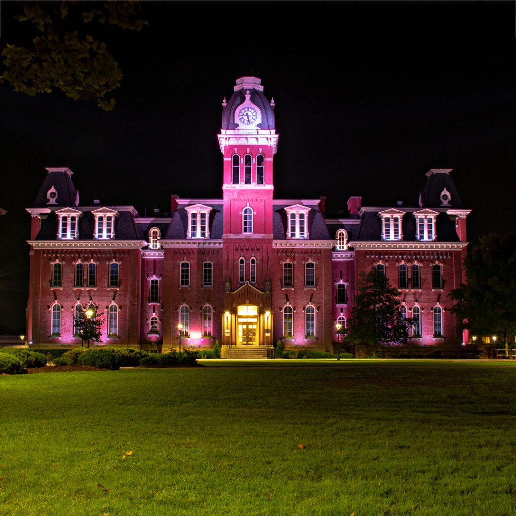 A historic brick building with a central clock tower illuminated in pink lights at night. The structure is surrounded by a well-maintained lawn and trees. The lit entrance and detailed architecture stand out against the dark sky.