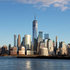 Skyline of a city with a prominent, tall skyscraper under a blue sky, viewed from across a body of water. Buildings of varying heights surround the central tower, and sunlight casts shadows on the architectural landscape.