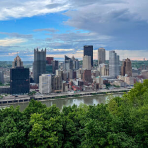 Panoramic view of a city skyline with modern skyscrapers, seen from across a river. The foreground features lush green trees, while the sky is partly cloudy with patches of blue.