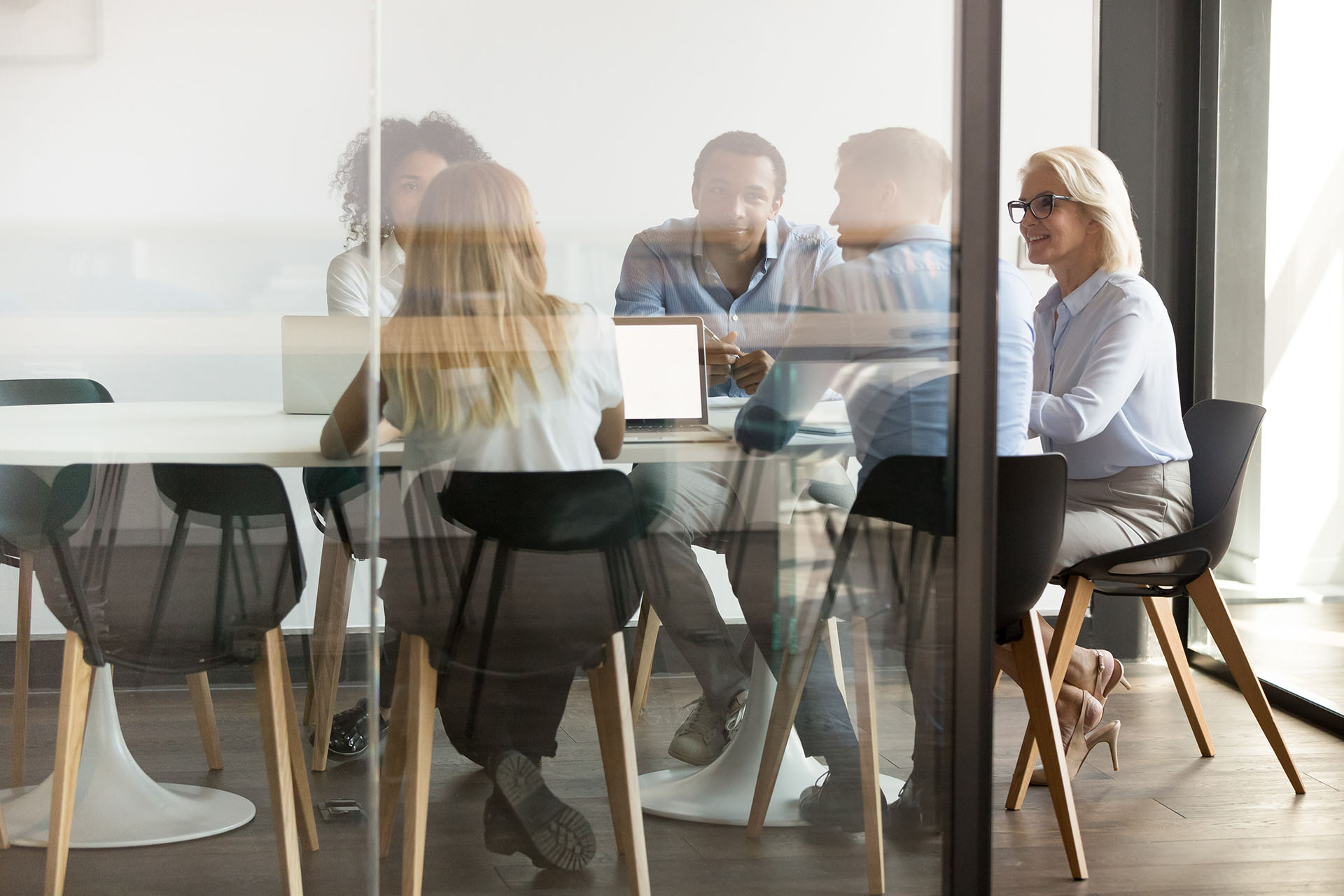 A group of five people in casual business attire sits around a conference table with laptops in a glass-walled meeting room. Engaged in a discussion about web design, one person takes notes while the others listen attentively.