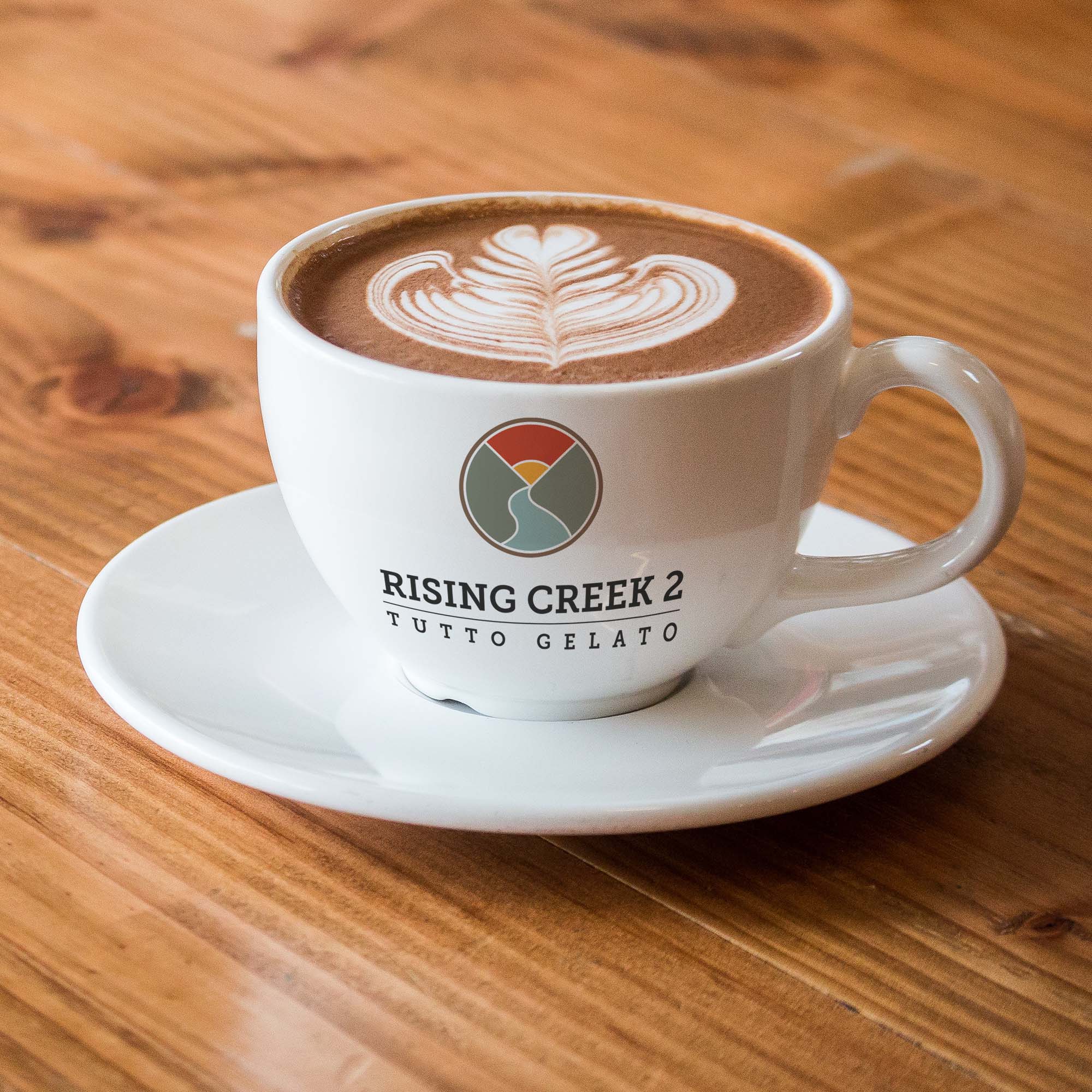 A latte with intricate foam art is served in a white cup and saucer, featuring a logo for "Rising Creek 2 Tutto Gelato." The cup is placed on a light wooden table.