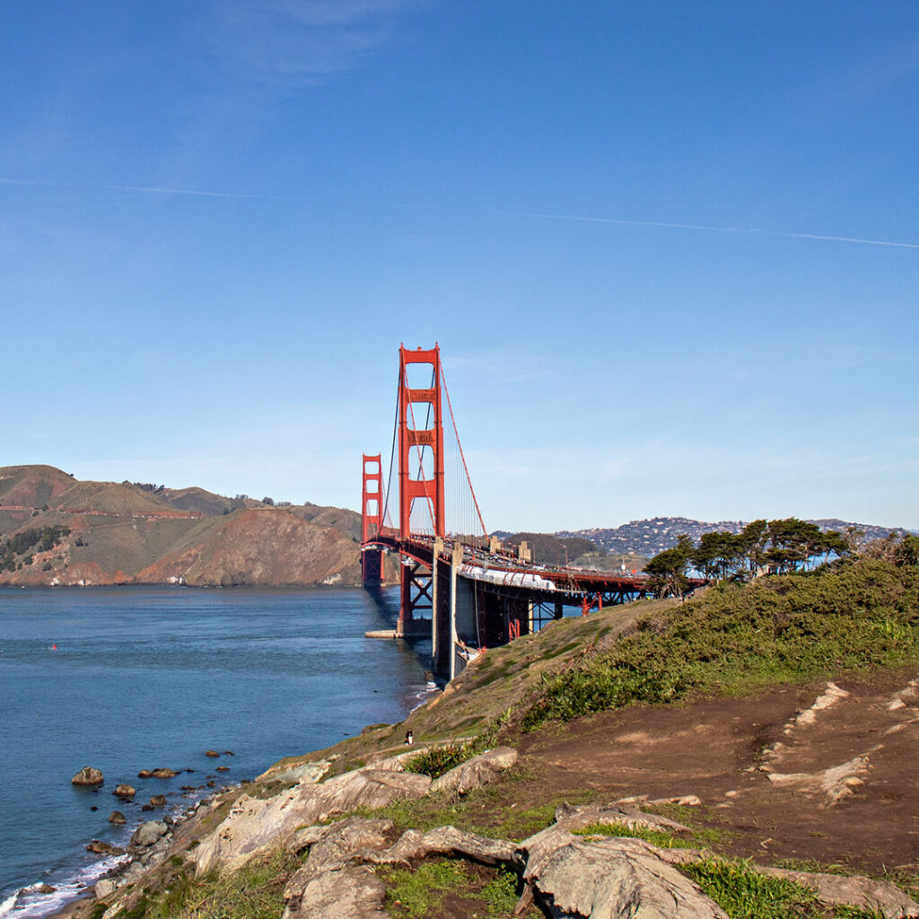 The image shows the Golden Gate Bridge stretching across the water, with bright red towers standing against a clear blue sky. Rocky shorelines and green hillsides are visible in the foreground and background.