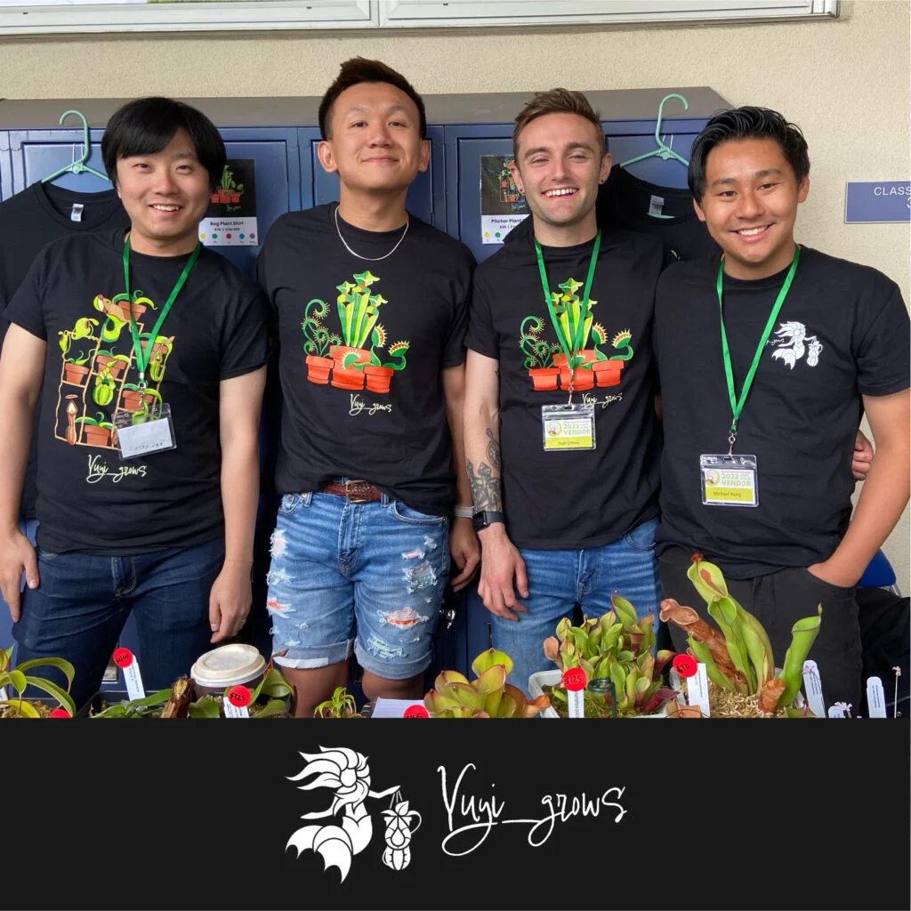 Four people stand together, smiling, wearing black "Kuii_grows" t-shirts with plant designs. They are in front of a wall with lockers and a display of small potted plants in front of them.