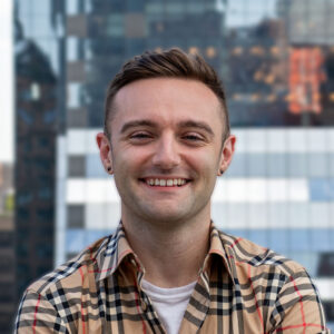 A smiling person with short hair and earrings stands outdoors against a blurred urban backdrop of modern glass buildings. They are wearing a checkered shirt over a white top.