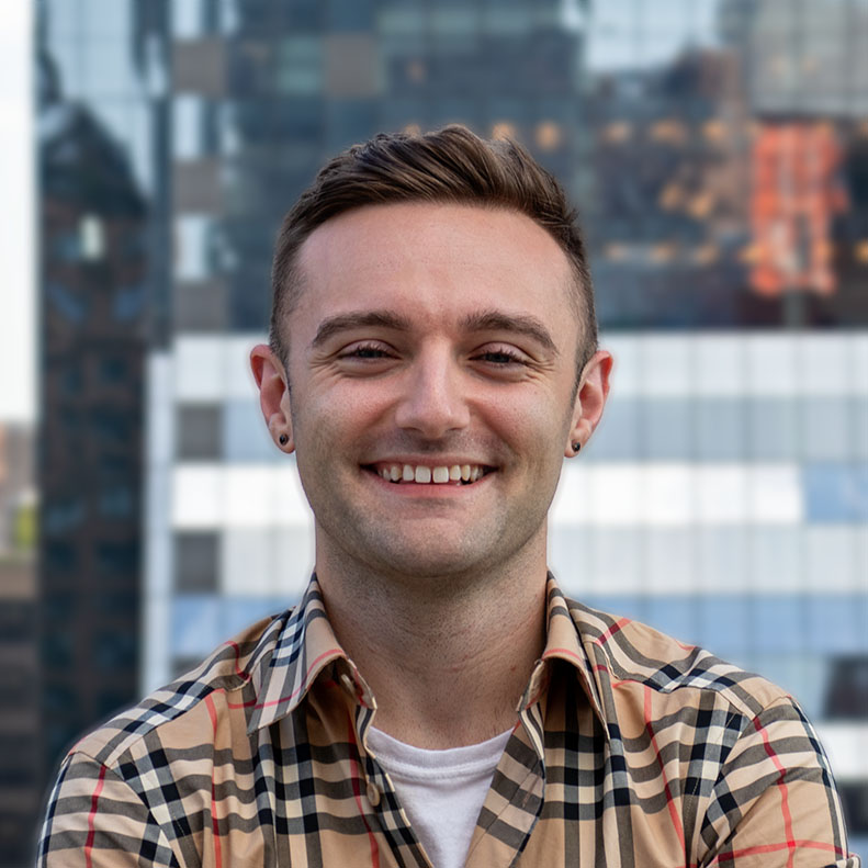 A smiling person with short hair and earrings stands outdoors against a blurred urban backdrop of modern glass buildings. They are wearing a checkered shirt over a white top.