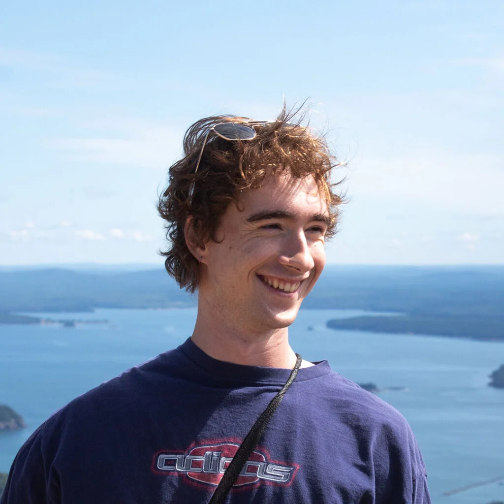 A person with curly hair is smiling against a backdrop of a vast body of water and distant hills under a clear blue sky. They are wearing a dark blue shirt with the visible text "adi-." The image conveys a sense of happiness and being outdoors.