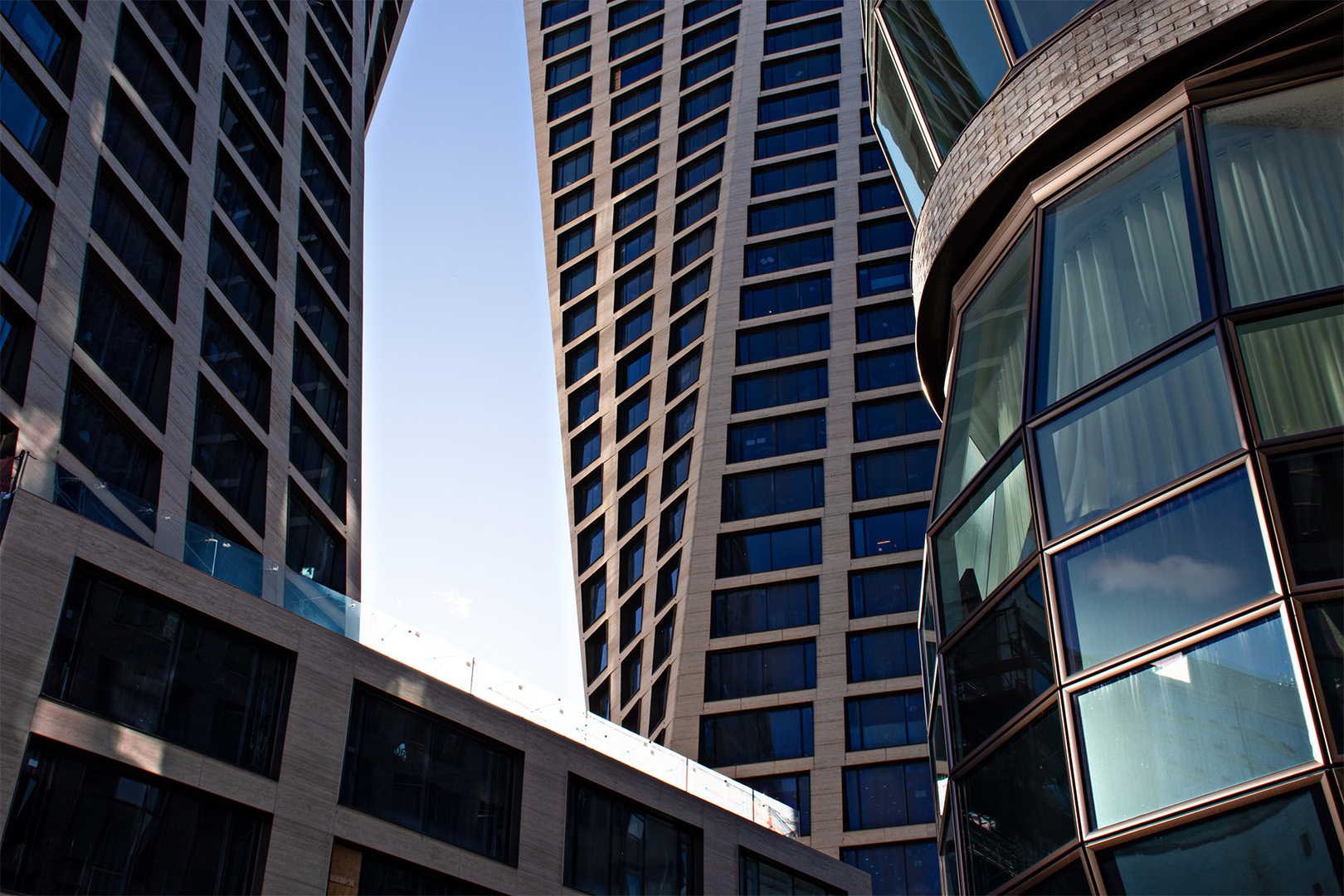 A view of modern architecture with three tall, twisting buildings set against a clear blue sky. Each structure features numerous windows, reflecting the sunlight and creating geometric patterns. The perspective emphasizes the buildings' unique angles.